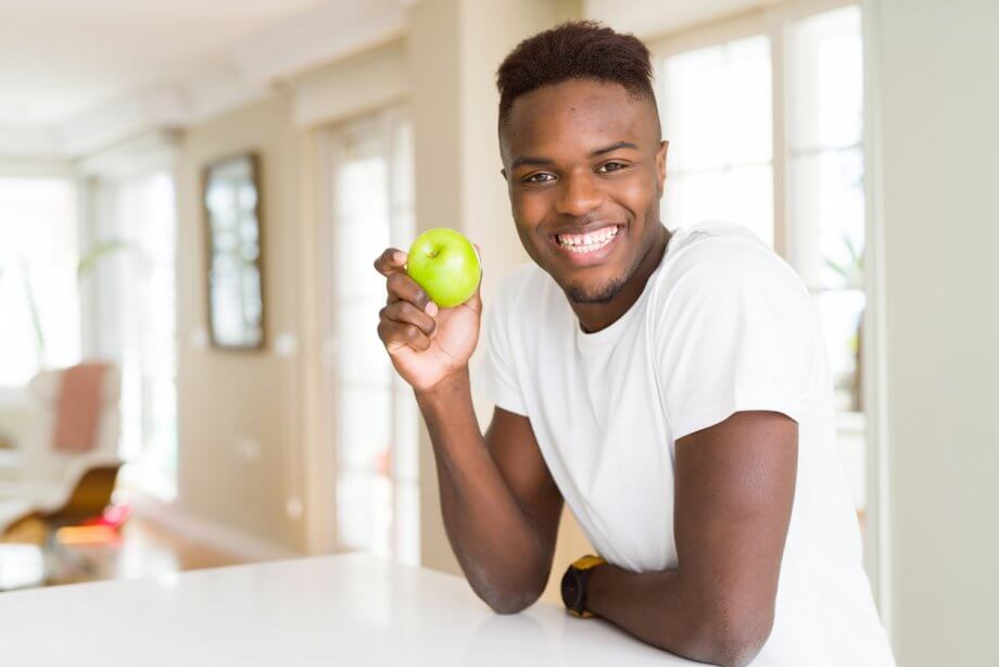 Young man with short dark hair is holding a green apple and smiling while in a kitchen.