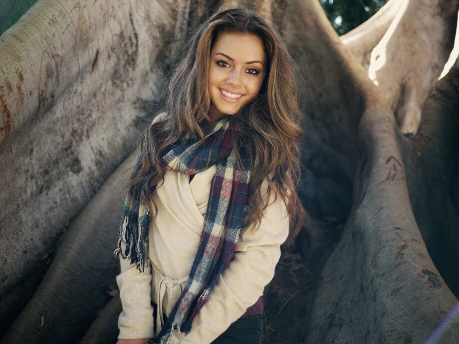 Young woman with long straight brown hair wearing a jacket and scarf is smiling while standing in front of a large tree.